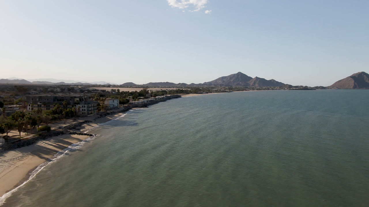 Aerial frame in San Felipe, Mexico, showing shoreline buildings closer to the left edge, shallow waves along the beach, broad green-blue water, hazy mountains, and no visible people.