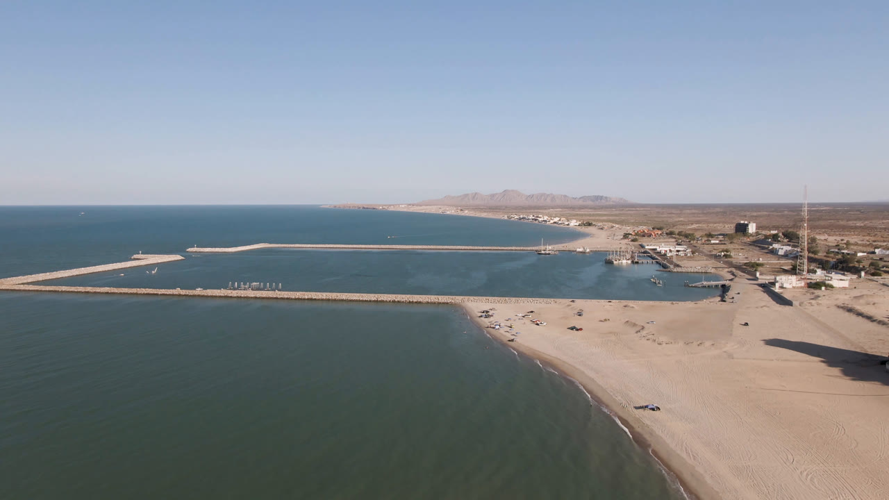 Opening aerial frame over San Felipe, Mexico showing green-blue water, a wide sandy beach, long breakwaters, distant mountains, shoreline buildings, and no visible people.