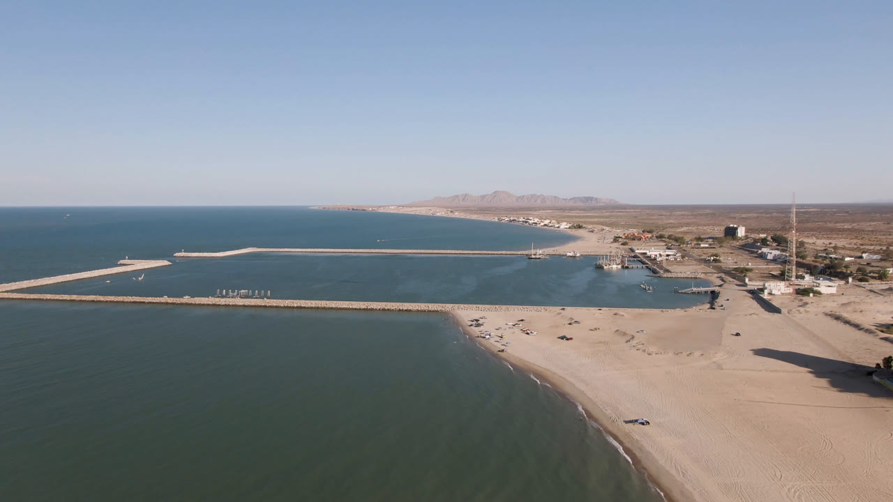 Aerial frame over San Felipe, Mexico with the beach curving along green-blue water, harbor structures extending across the bay, low buildings onshore, mountains on the horizon, and no visible people.