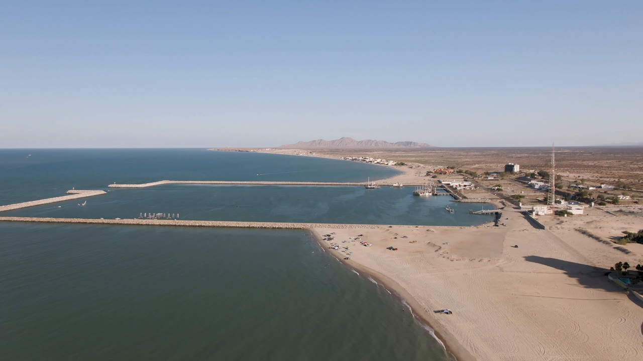 San Felipe, Mexico aerial view with a broad sandy beach below, green-blue water inside the breakwaters, small boats near the harbor, a tall mast or tower onshore, and no visible people.