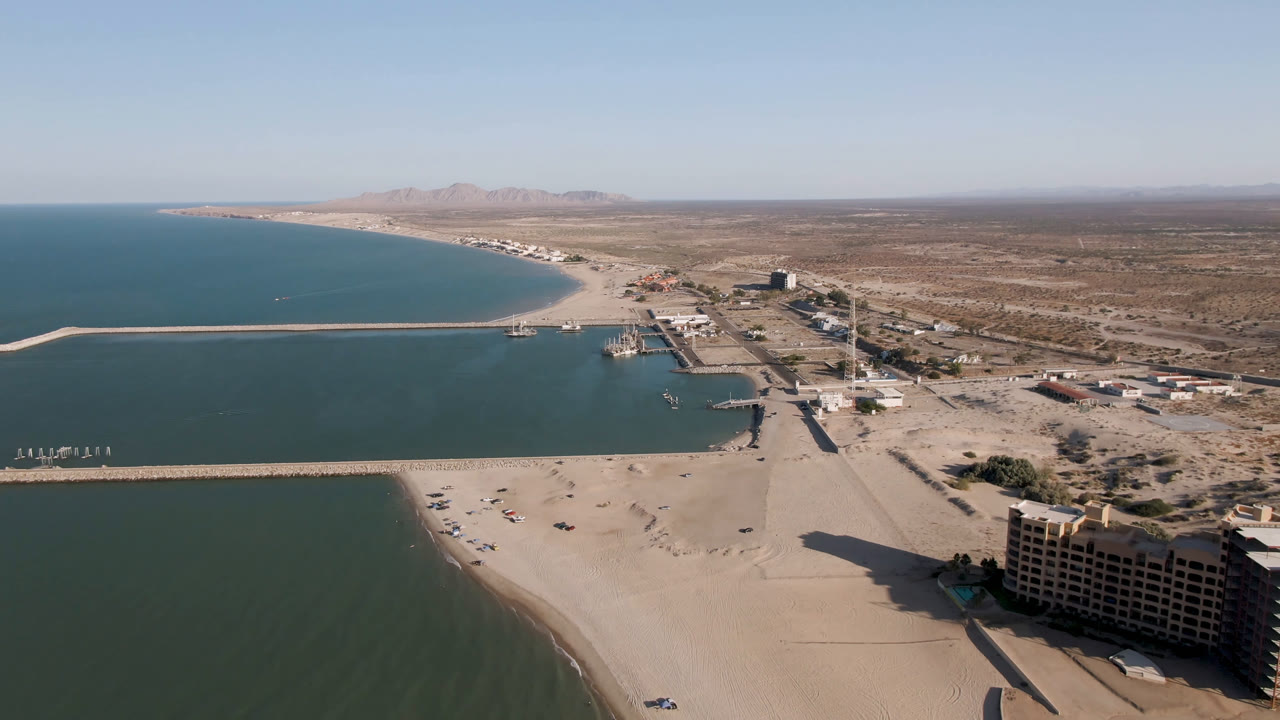 Wide aerial frame in San Felipe, Baja California, showing the curved shoreline, pier crossing the water, scattered buildings, desert flats, and no visible people.