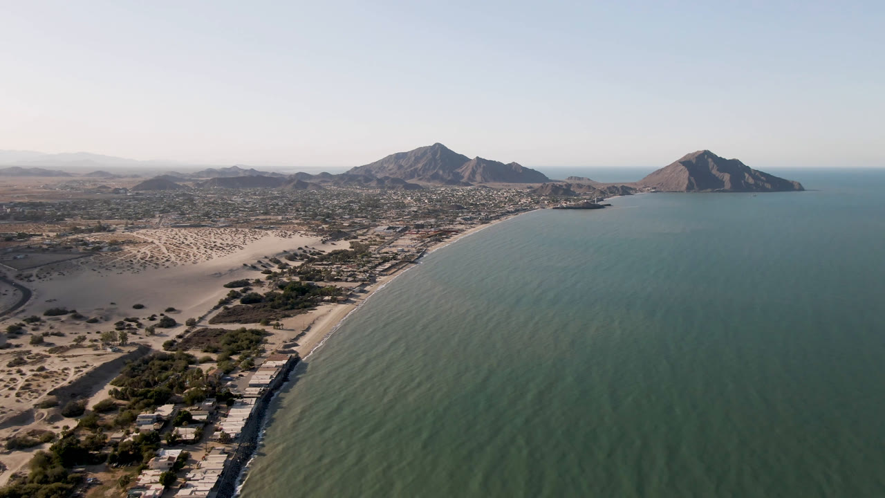 Drone-like aerial of coastline with ocean, beach, desert settlement, and mountains.
