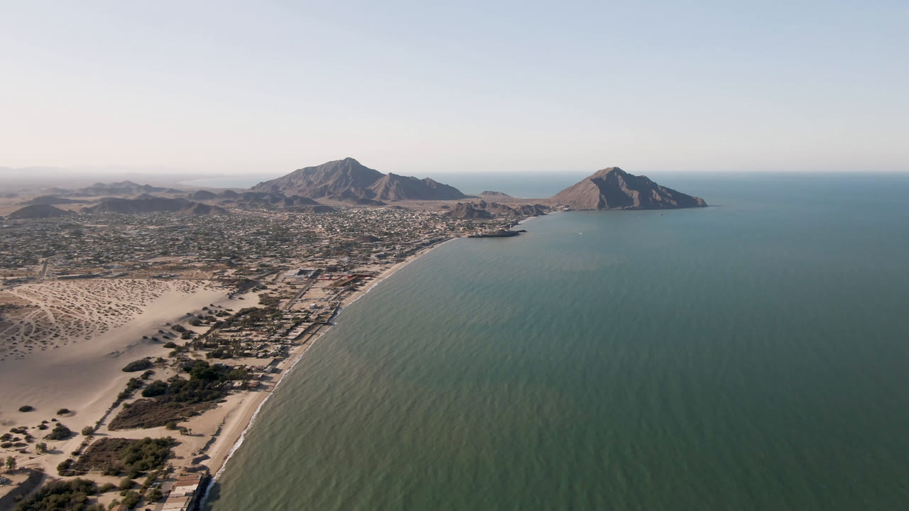 Wide aerial frame over San Felipe, Mexico, showing a curved sandy beach, green-blue bay water, low town grid, rocky mountains, clear sky, and no visible people.
