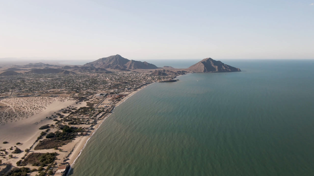 Aerial view of San Felipe, Mexico, with pale dunes at lower left, a long curved shoreline, green-blue water, low buildings inland, distant mountains, and no visible people.