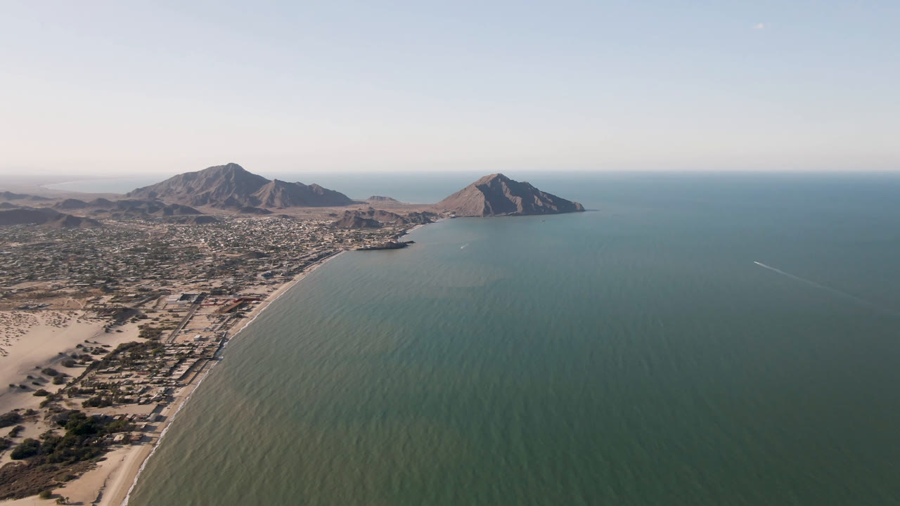 Wide coastal aerial with beach, town, bay water, and mountain backdrop