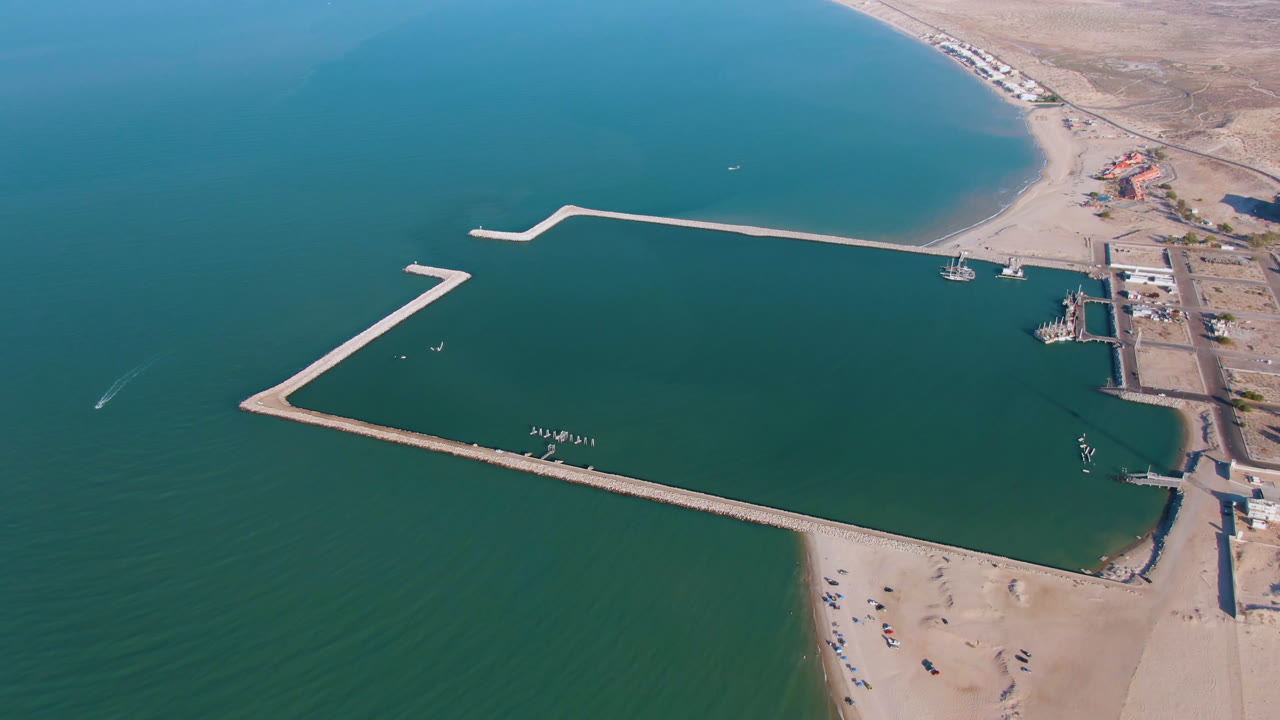 Opening aerial frame over San Felipe, Baja California, showing blue-green water, angular pale breakwaters, sandy beach, shoreline road, low buildings, dry land, and no visible people.