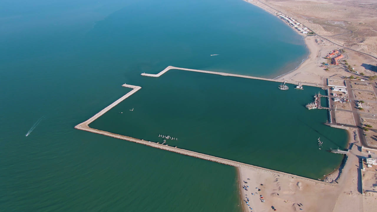 Aerial view over San Felipe, Baja California, with blue-green water, a sandy beach, an angular breakwater, shoreline buildings, desert terrain, and no visible people.