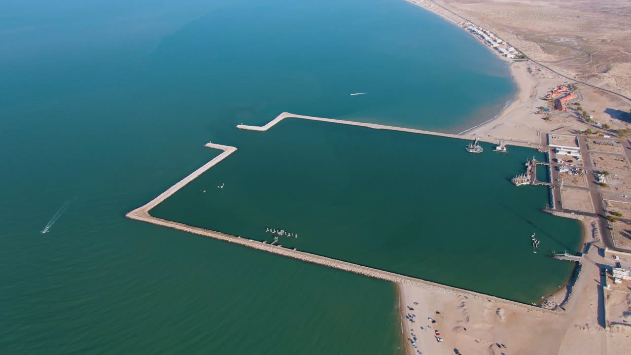 High aerial frame in San Felipe, Baja California, showing the long breakwater enclosing water, a sandy shoreline below, small coastal structures, arid land beyond, and no visible people.