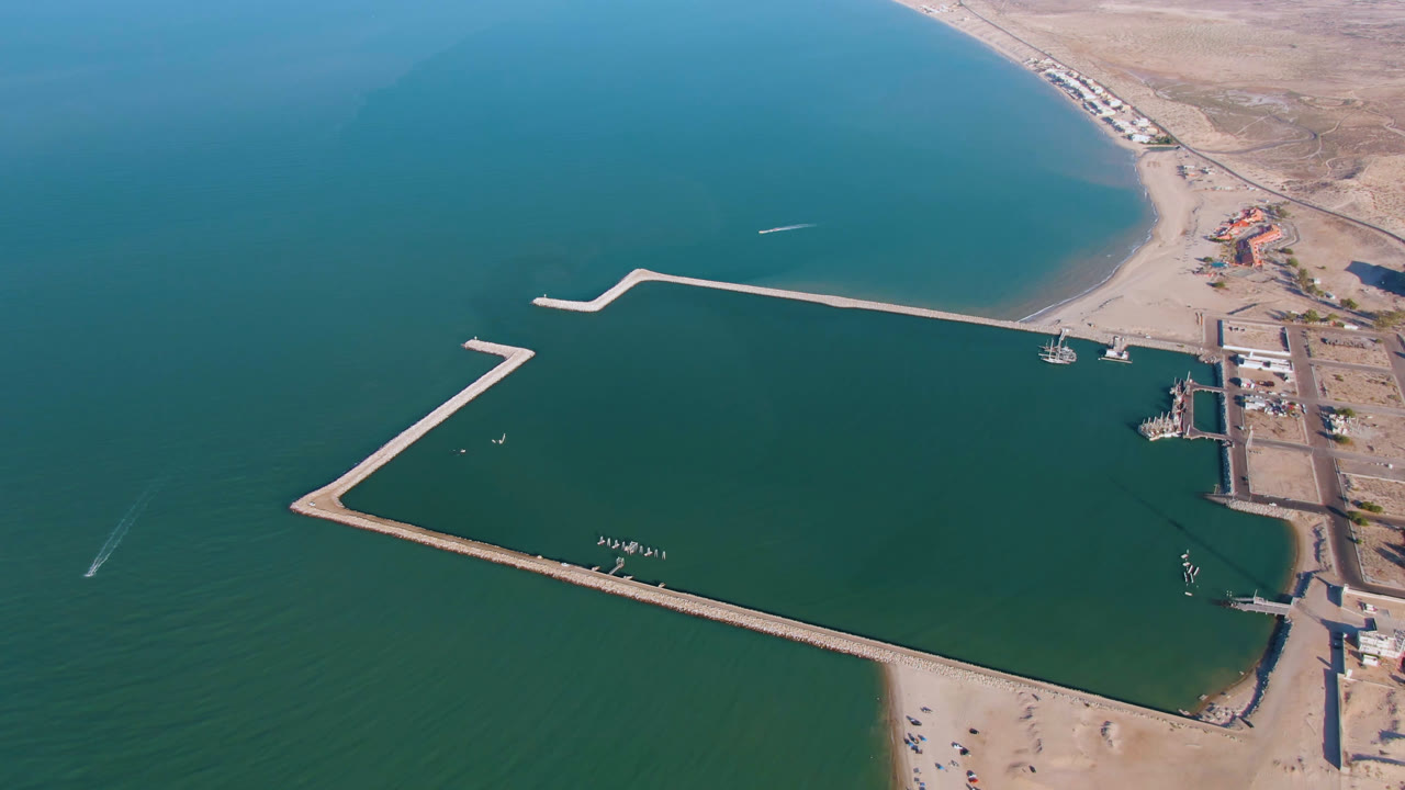 Aerial frame over San Felipe BC, Mexico showing a long angular dock enclosing teal water, sandy shoreline below, dry inland terrain at right, and no visible people.
