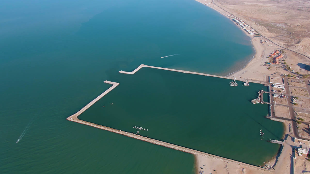 Aerial view over San Felipe coastal water with an angular breakwater, sandy beach, dry terrain, and no visible people at the start of the clip.