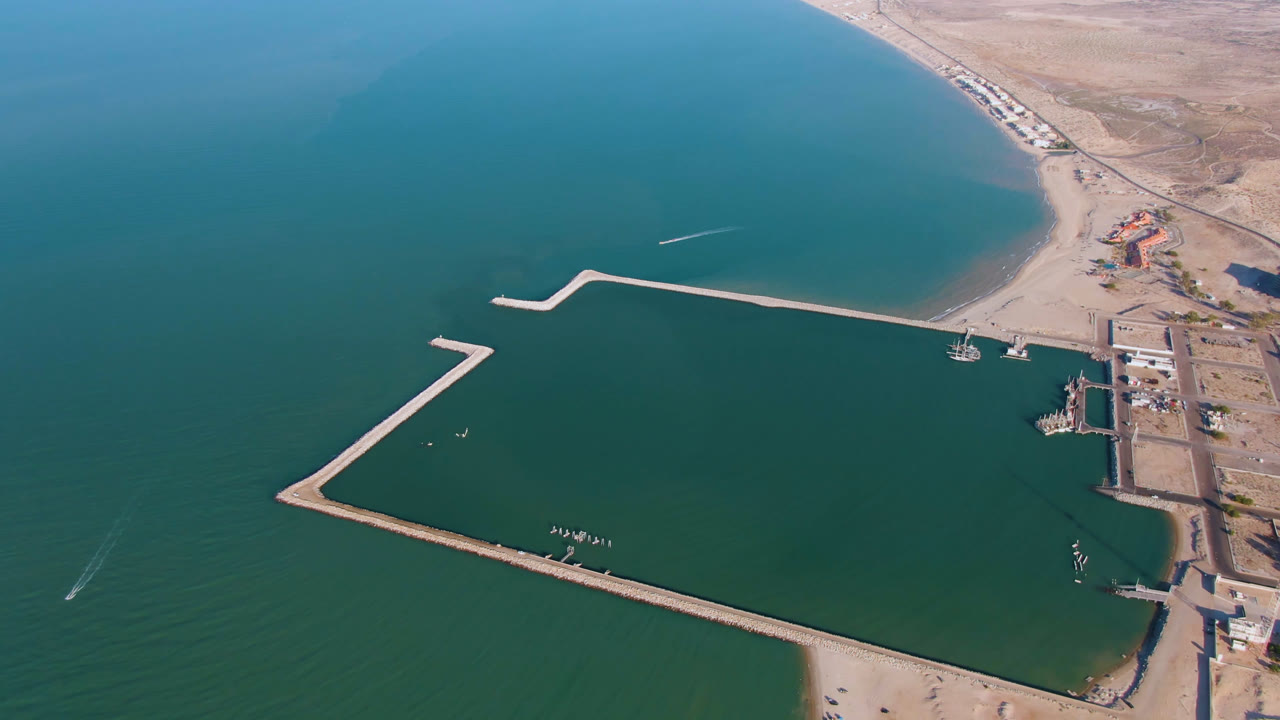 High aerial frame of San Felipe with the breakwater spanning the water, a dock area along shore, pale desert land behind it, and no visible people.