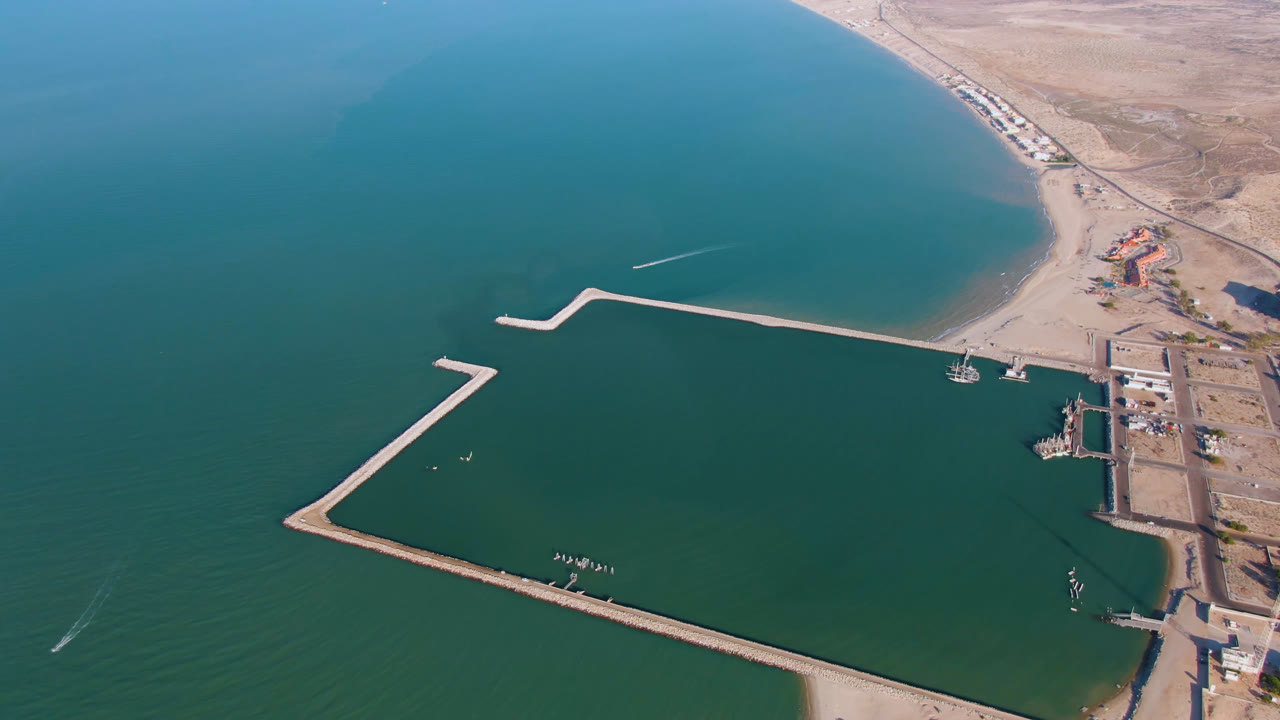 High aerial frame over San Felipe, Baja California, Mexico, showing teal water, a pale beach, a long angular breakwater, dock structures at right, and no visible people.