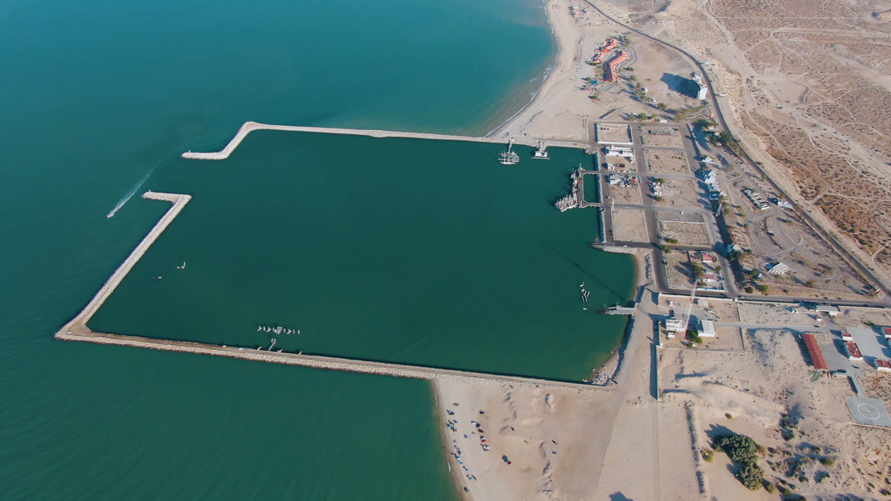 Overhead aerial frame from San Felipe, Baja California, Mexico showing teal water around pale harbor breakwaters, a sandy beach at the bottom, dry shoreline on the right, and no visible people.