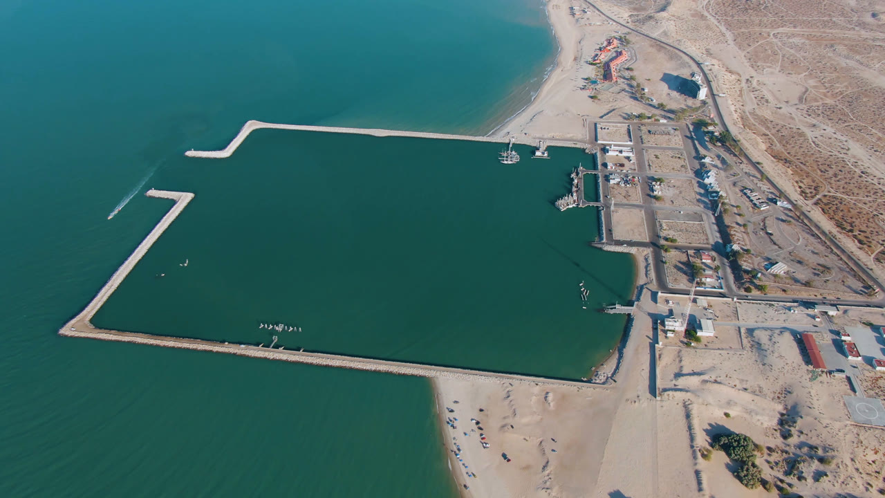 High drone view over San Felipe, Baja California, Mexico with the long pale breakwater crossing the lower water, docks near the right shoreline, sandy beach below, and no visible people.