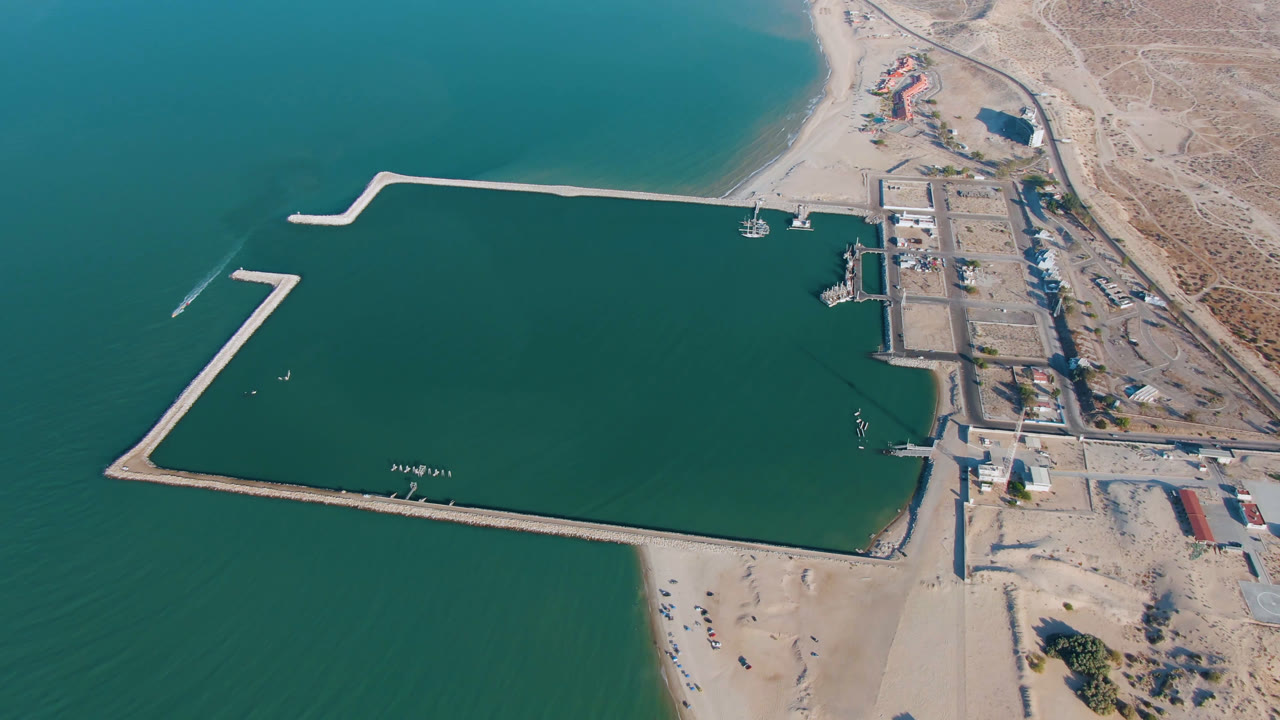Drone view of a coastal harbor with green-blue water, breakwaters, docks, beach, and arid land.