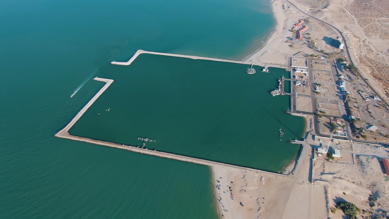 Aerial frame over San Felipe, Baja California showing teal water enclosed by pale breakwaters, a sandy beach at the bottom, dock structures at right, desert ground beyond, and no visible people.
