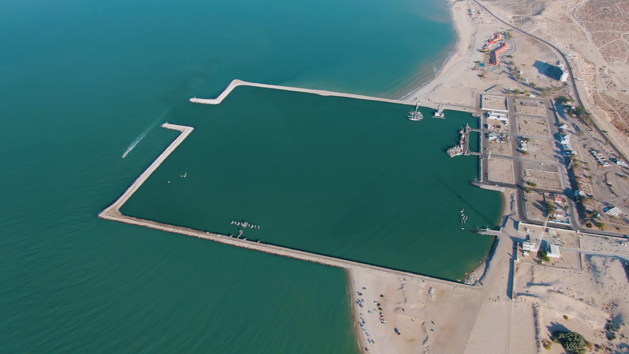 Aerial frame over San Felipe, Baja California with the long lower breakwater crossing the water, the upper breakwater reaching from shore, small watercraft near the dock, sandy beach below, and no visible people.