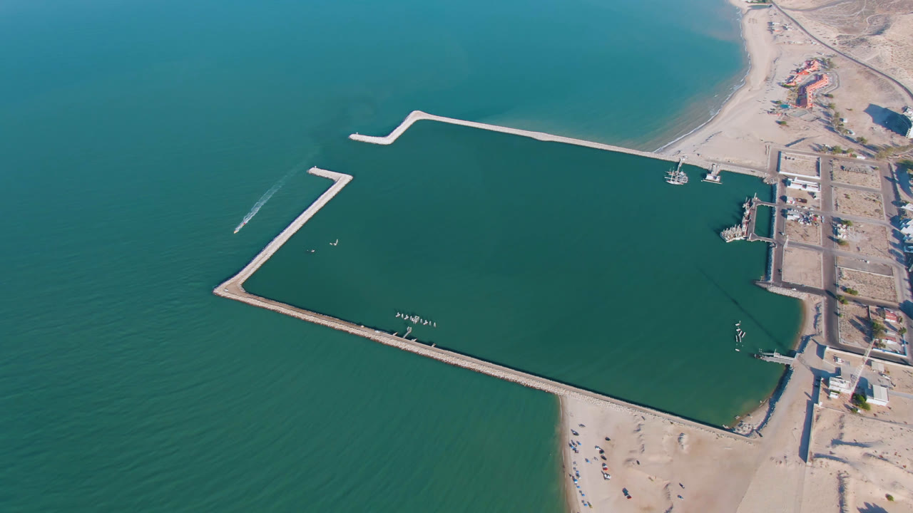 Opening aerial frame over San Felipe, Mexico, showing turquoise water around pale breakwaters, a sandy beach at the bottom edge, shoreline buildings at right, and no visible people.