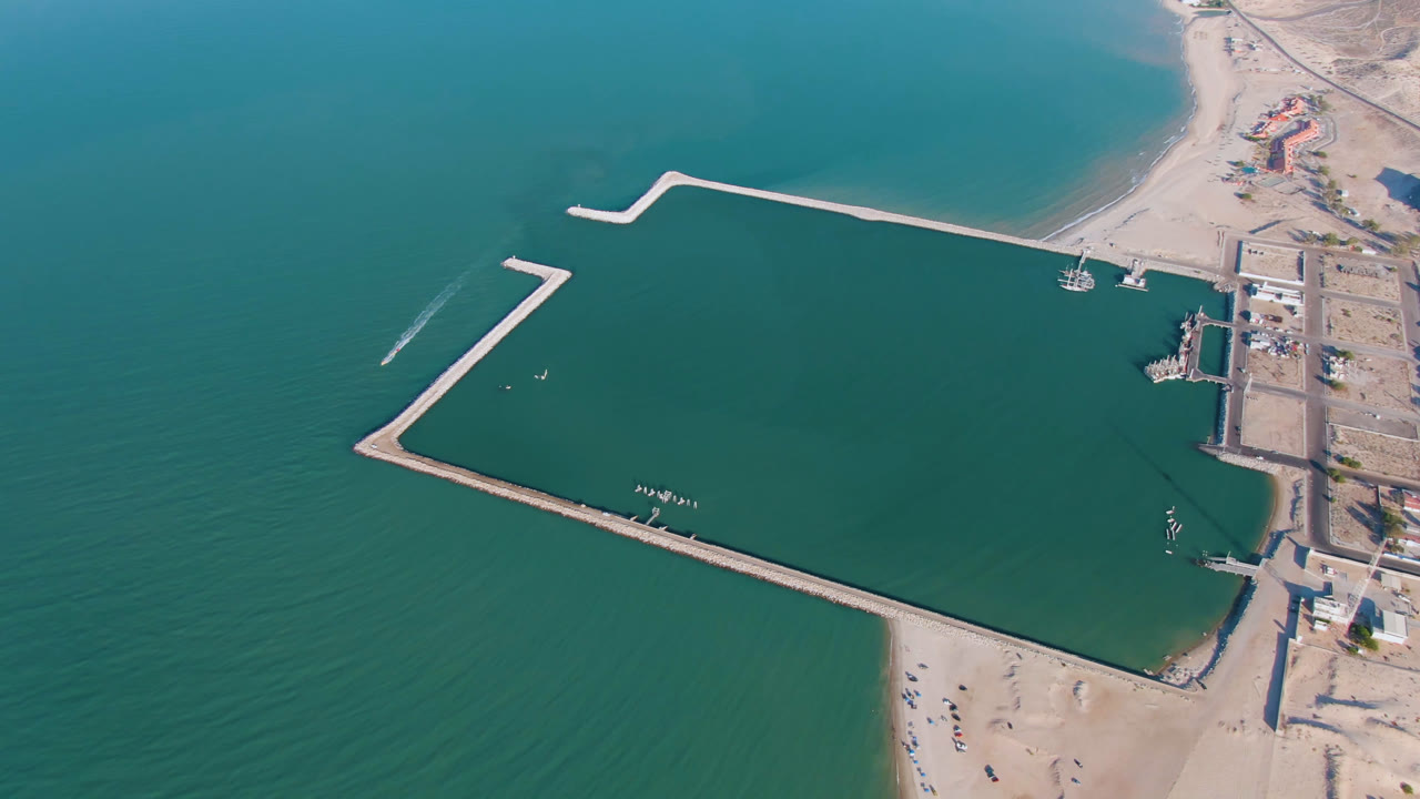 Aerial frame over San Felipe, Mexico, with the lower breakwater stretching across green water, small boats near the harbor interior, beach below, and no visible people.
