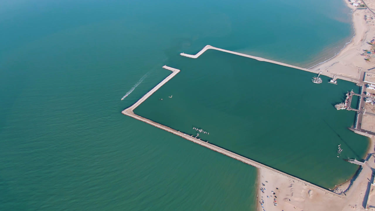 Opening aerial frame over San Felipe water with angular pale breakwaters, sandy beach at the lower edge, small boats near the wall, and no visible people.