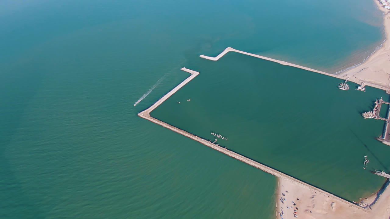 Drone view of open water, angular breakwaters, and a sandy beach corner.