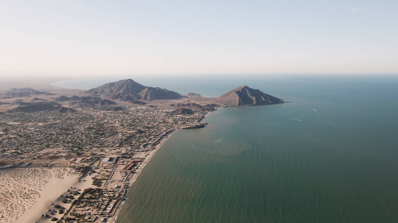 High aerial view over San Felipe, Mexico, with sandy beach at lower left, a coastal town beside green-blue water, mountains on the horizon, and no visible people.
