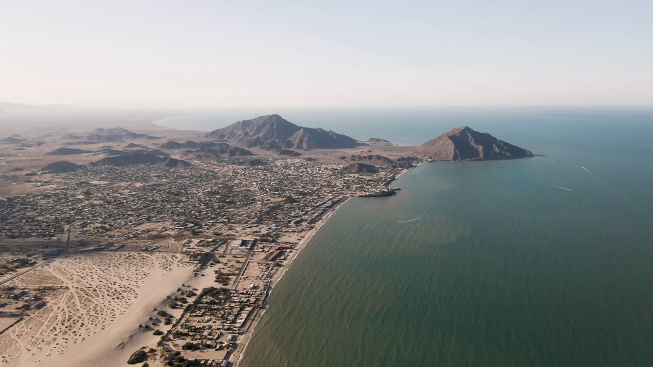 Aerial frame of San Felipe coastline with the beach and road network below, the city spread across the midground, mountains beyond the bay, and no visible people.