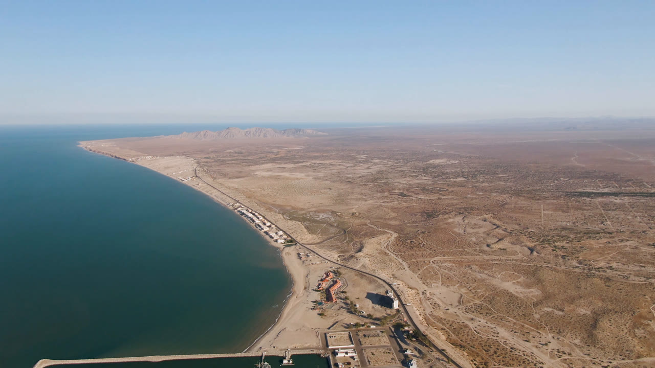 Opening aerial frame over San Felipe, Baja California, Mexico, with blue-green water on the left, a sandy beach curving through the center, dry desert on the right, distant mountains near the horizon, and no visible people.