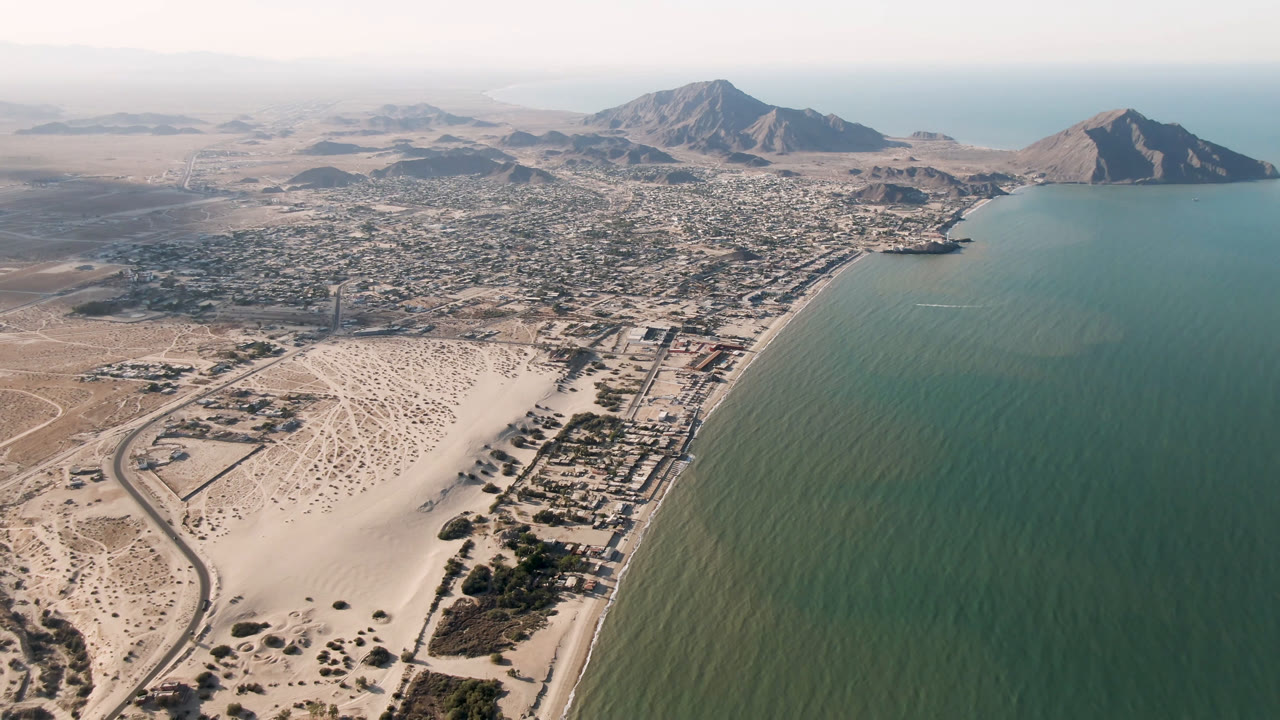 Opening aerial frame over San Felipe, Baja California, Mexico, showing green coastal water, pale sand, desert roads, low city blocks, distant mountains, and no visible people.