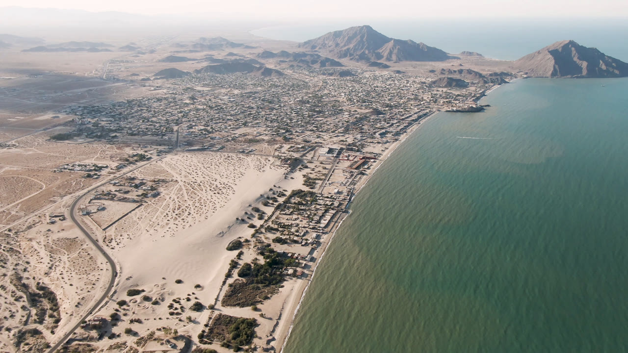 Wide aerial of coastal city beside green sea with pale beach and mountains beyond.