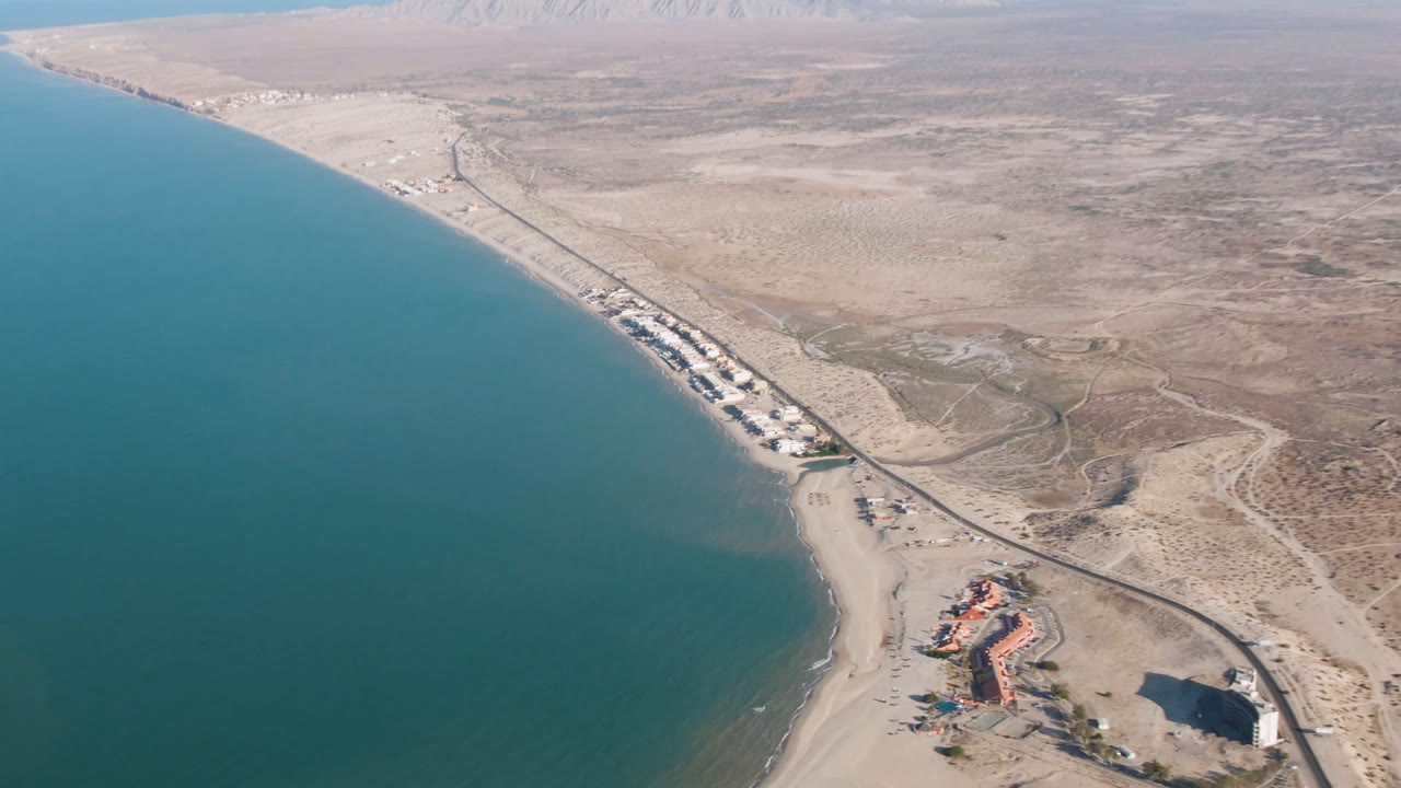 Drone view of coast with water, sand, road, scattered buildings, and mountains in the distance.