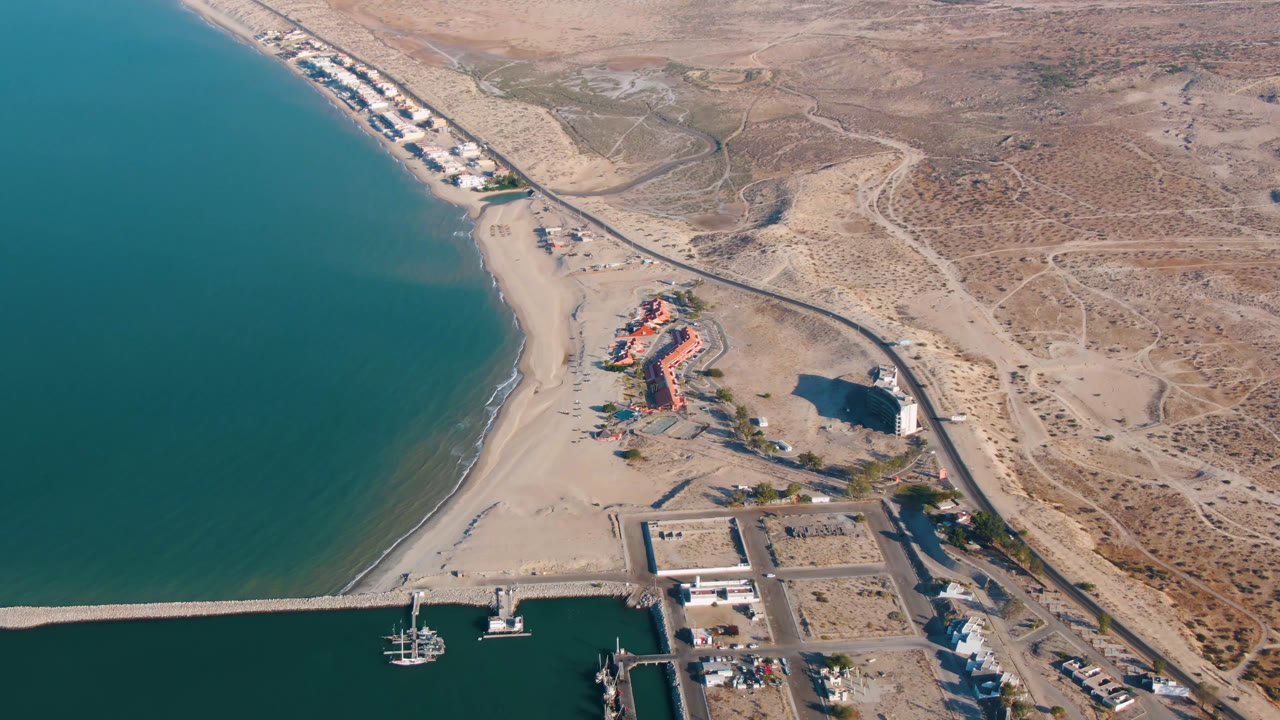 Opening aerial frame over San Felipe, Mexico with teal water at left, a sandy beach, a harbor breakwater at the bottom, a long road, scattered buildings, dry inland terrain, and no visible people.
