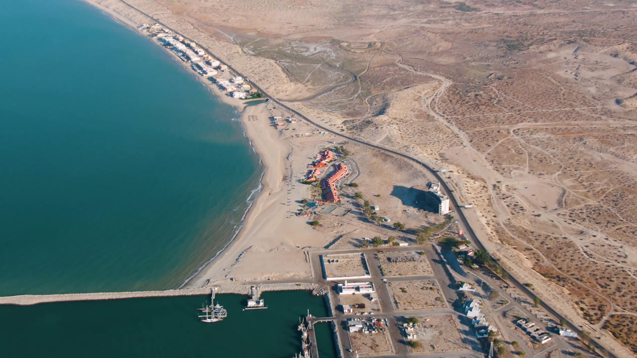 Aerial frame over San Felipe, Mexico showing the shoreline slightly farther along, with the harbor breakwater across the lower edge, boats near the docks, beach beside teal water, a long road, and no visible people.