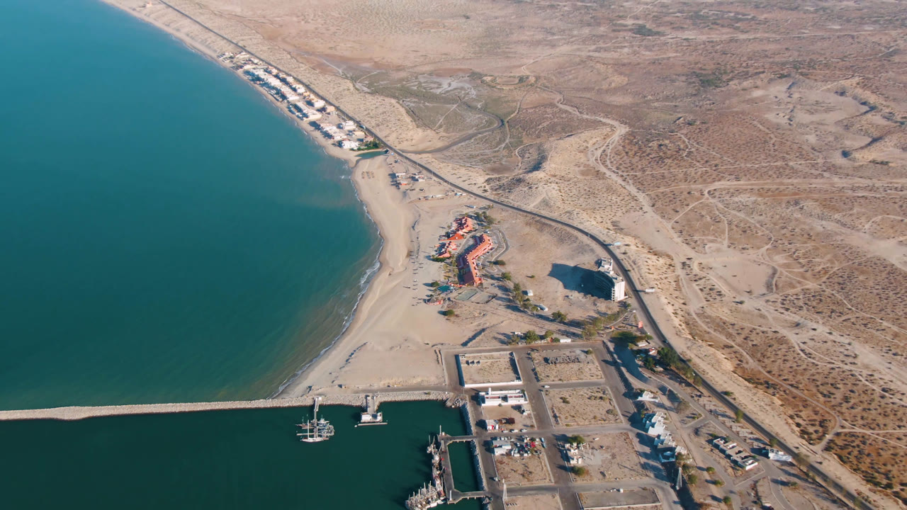 Drone view of coastline with beach, road, harbor area, boats, buildings, and desert beyond.