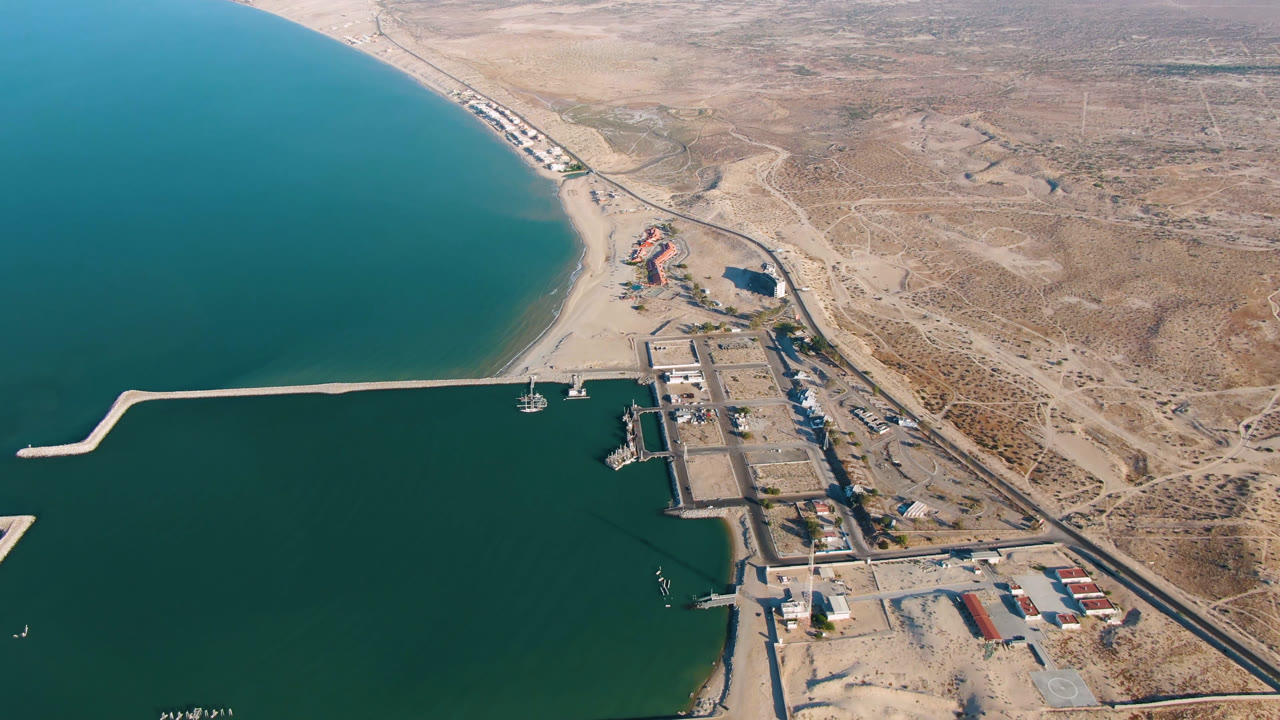Opening aerial frame over San Felipe BC Mexico showing teal coastal water, a sandy beach, a long pale pier or breakwater, shoreline buildings, road, dry terrain, and no visible people.