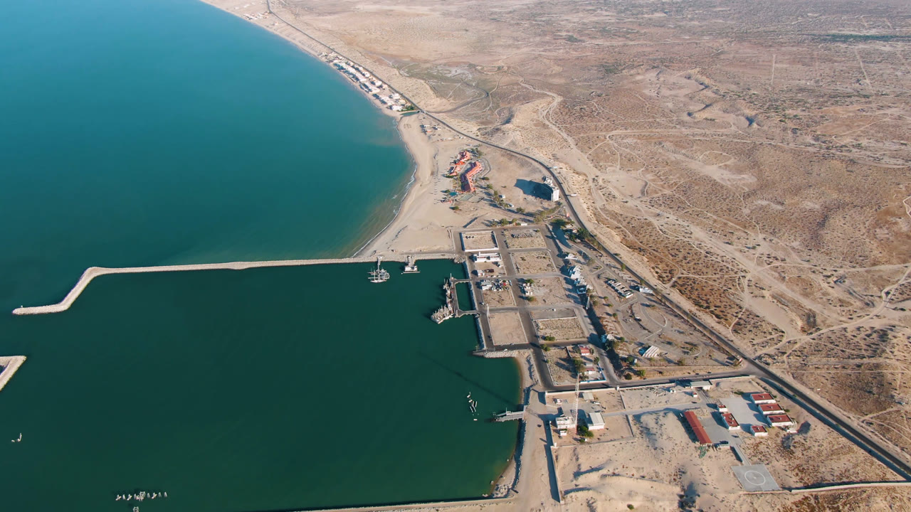 Second aerial frame over San Felipe BC Mexico with the long pier reaching across dark teal water, beach and harbor structures near center, road along the right, desert terrain beyond, and no visible people.