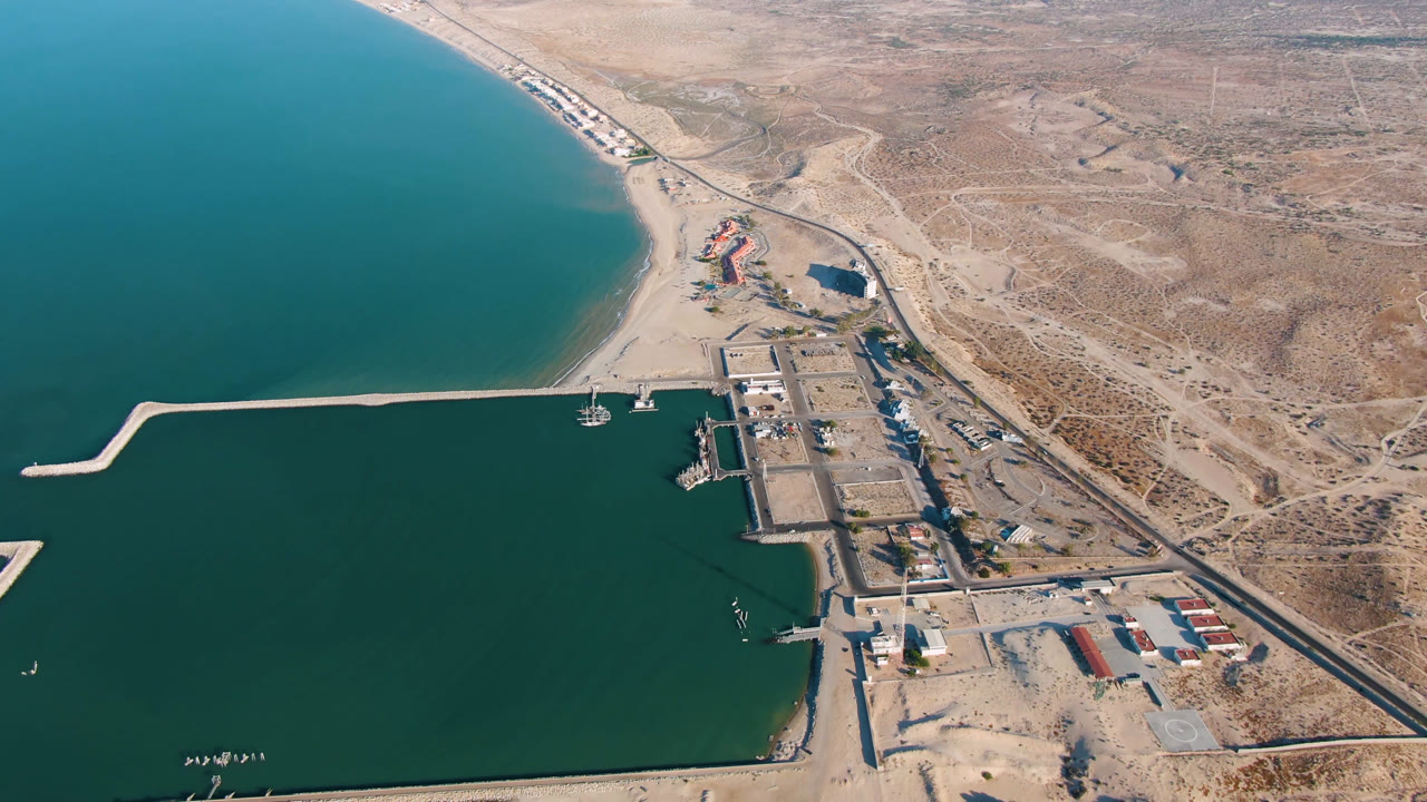 Aerial coastline view with pier, marina edge, beach buildings, road, and arid hills.