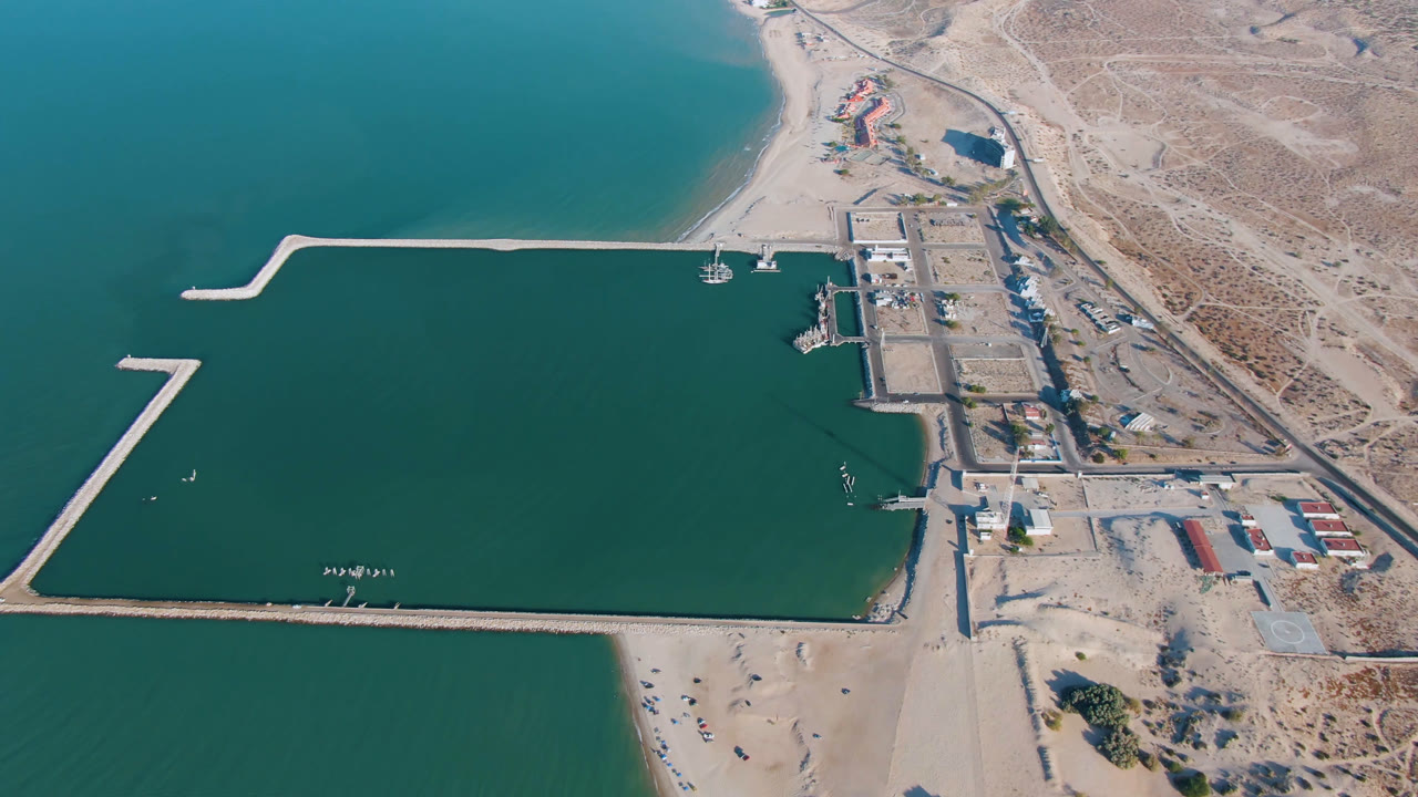 High aerial frame of San Felipe coastline with the breakwater stretching across teal water, beach at the lower edge, shoreline buildings to the right, and no visible people.