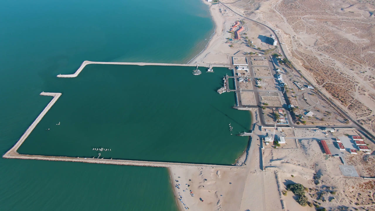 Aerial harbor scene with a long pier-like breakwater, docks, beach, and desert shoreline.