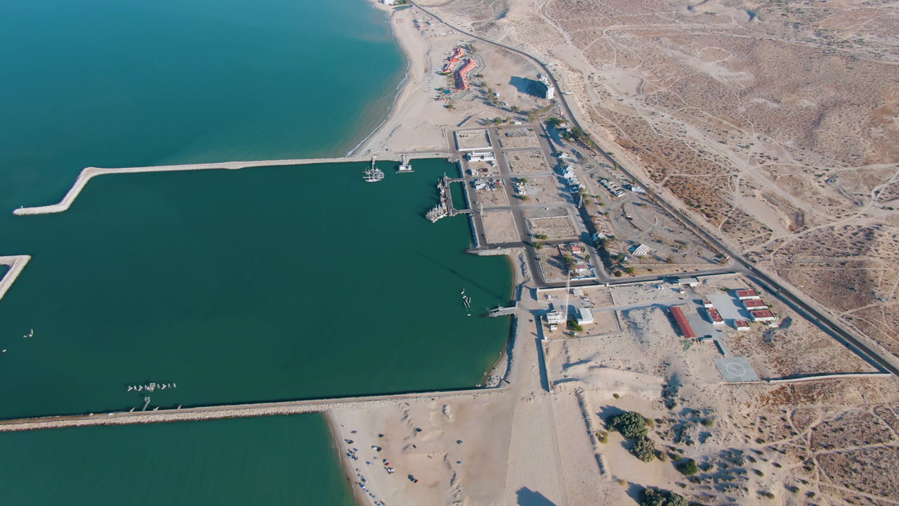 Opening aerial frame over San Felipe, Baja California, Mexico, showing teal water, harbor breakwaters, sandy beach, dock structures, a road, and dry terrain with no visible people.