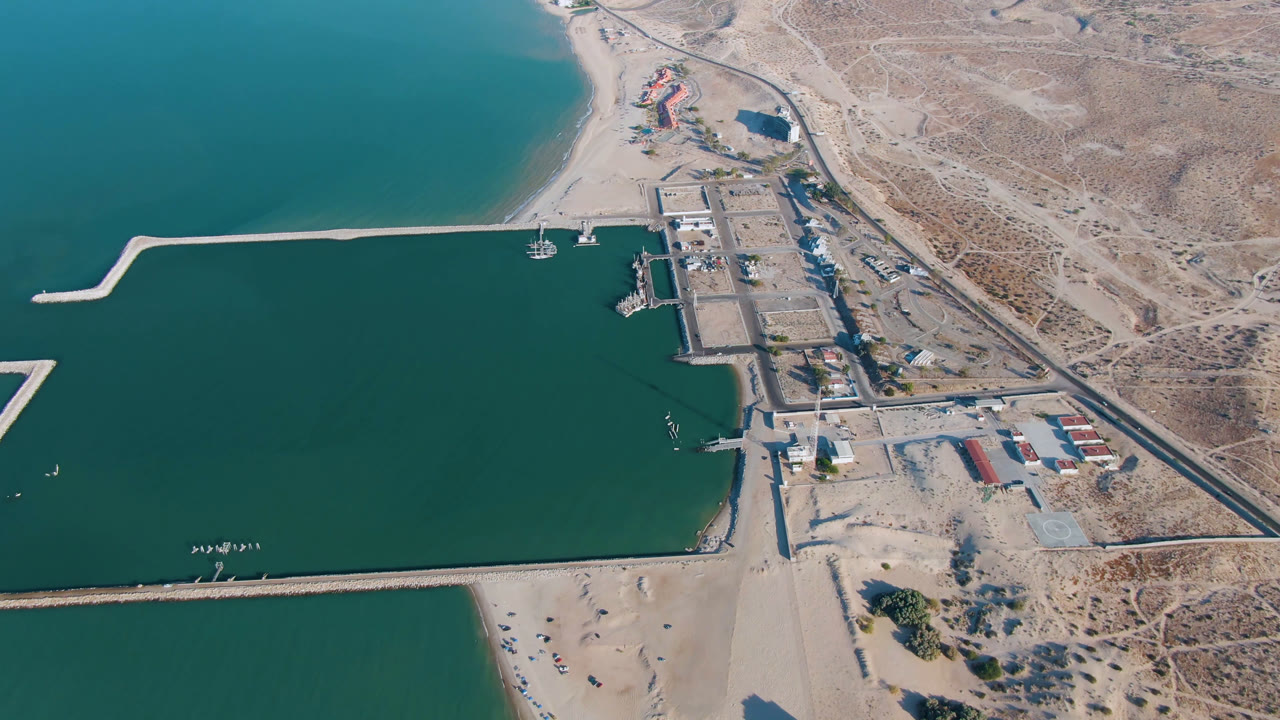High aerial of a harbor entrance, beach, dock area, road, and arid shoreline.
