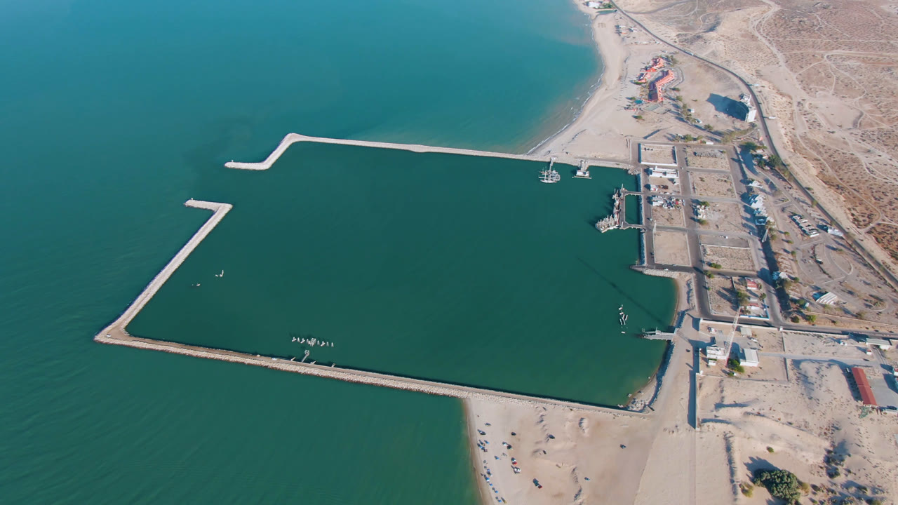 Opening aerial frame over San Felipe, Baja California, showing turquoise water, pale harbor walls, sandy beach, desert shoreline, small boats, and no visible people.