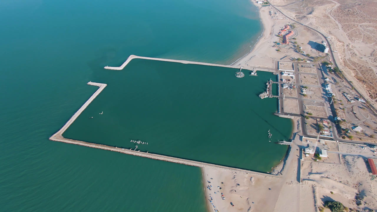 Aerial frame over San Felipe with the long outer breakwater centered, teal water surrounding the harbor, beach below, desert terrain to the right, and no visible people.