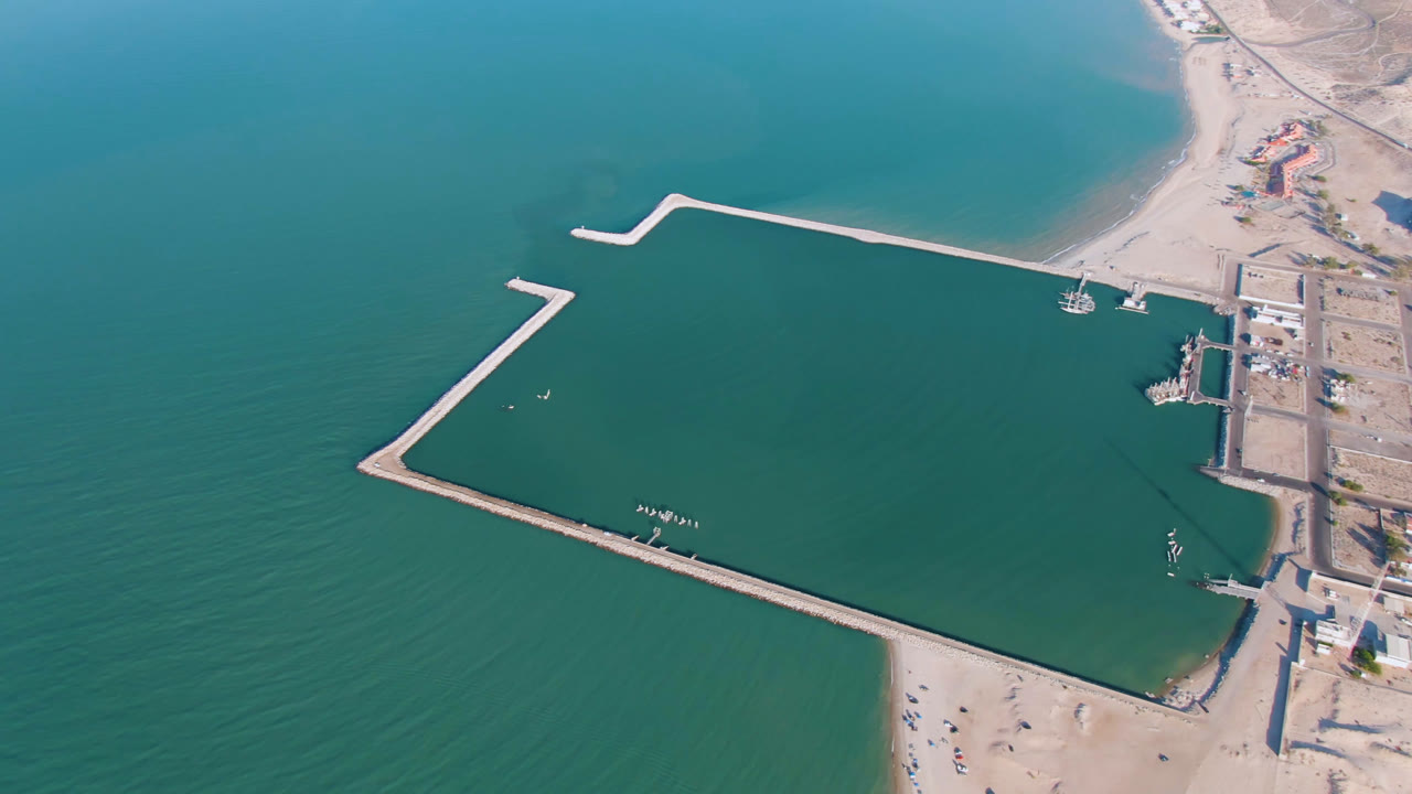 Opening aerial frame over San Felipe, Mexico, showing turquoise water, pale sand, dock structures, angular breakwaters, shoreline buildings, and no visible people.