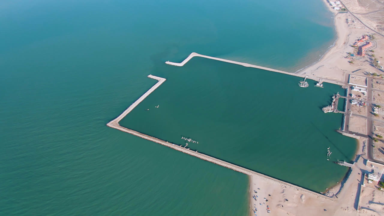 Aerial view over San Felipe, Mexico, with the lower breakwater stretching across the frame, a curved upper breakwater, small boats near the docks, and no visible people.