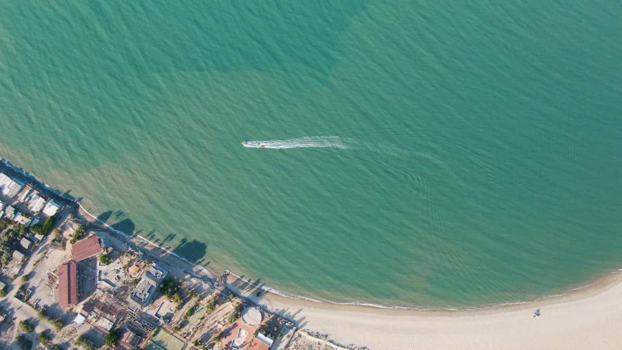 Overhead frame from San Felipe, Mexico showing a small white boat crossing green-blue water above a sandy beach and beachfront buildings, with no visible people.