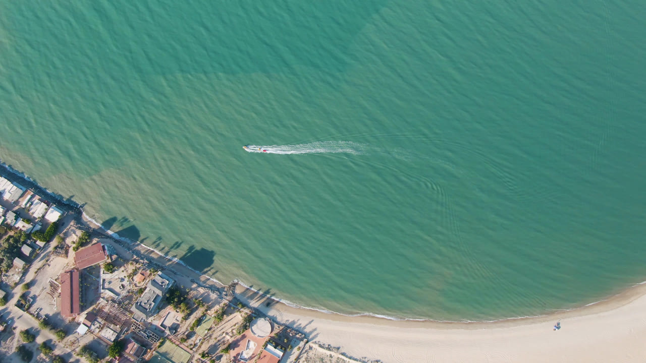 Overhead San Felipe frame with the boat farther across the water, a thin white wake behind it, pale sand below, and no visible people on the beach or boat.
