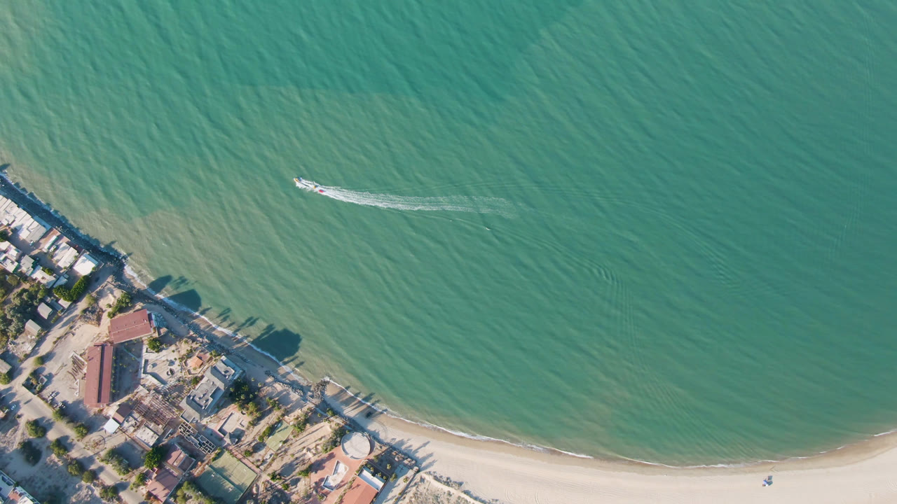 High aerial frame over San Felipe, Baja California, Mexico, with turquoise water filling the view, a sandy shoreline at the bottom, coastal buildings at lower left, a small boat near the upper center, and no visible people.
