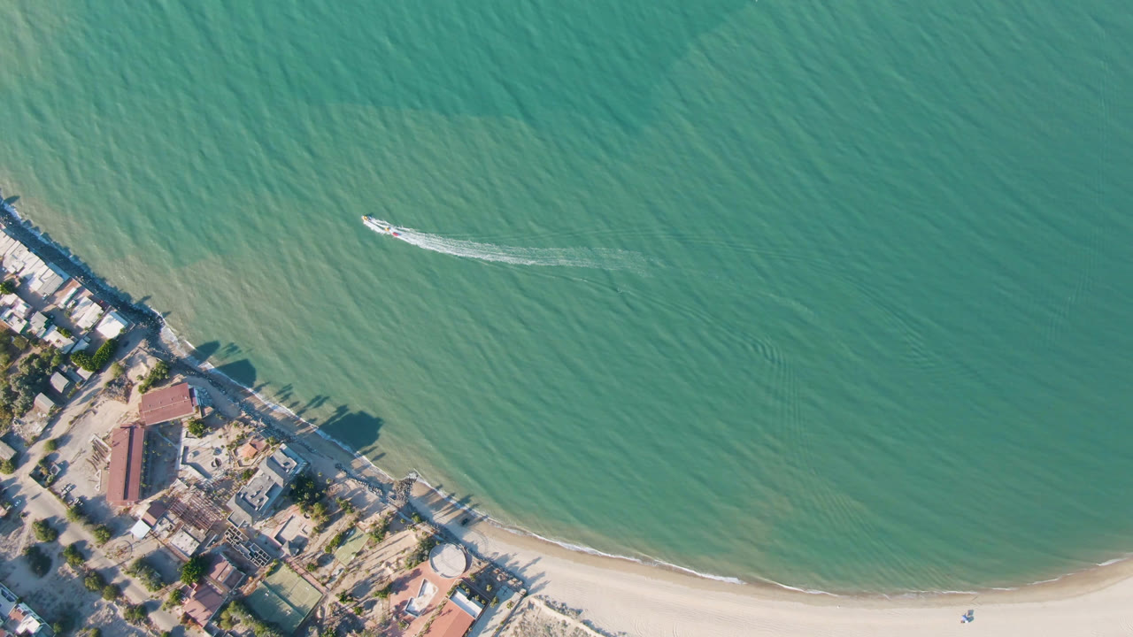Overhead San Felipe beach view with the boat slightly farther across the green-blue water, a thin white wake trailing behind it, sand along the lower edge, buildings and trees at lower left, and no visible people.