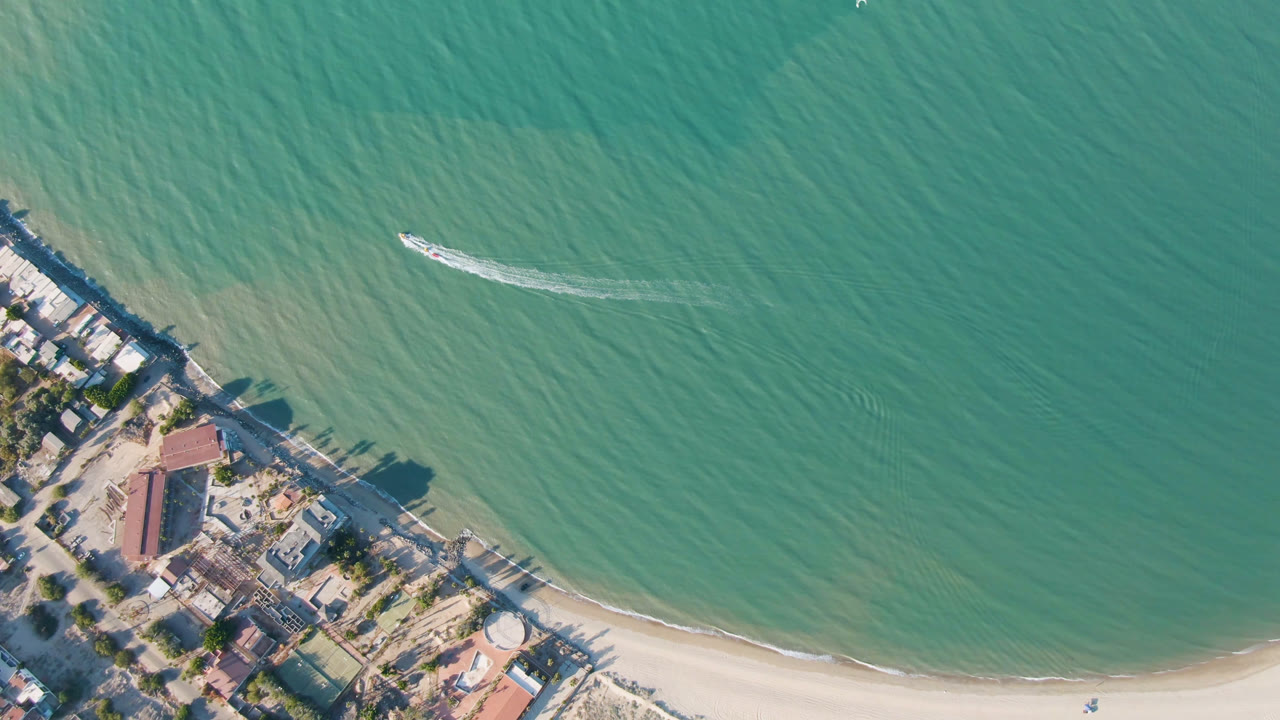 Aerial beach frame with a boat wake on turquoise water and buildings along the lower left shore.